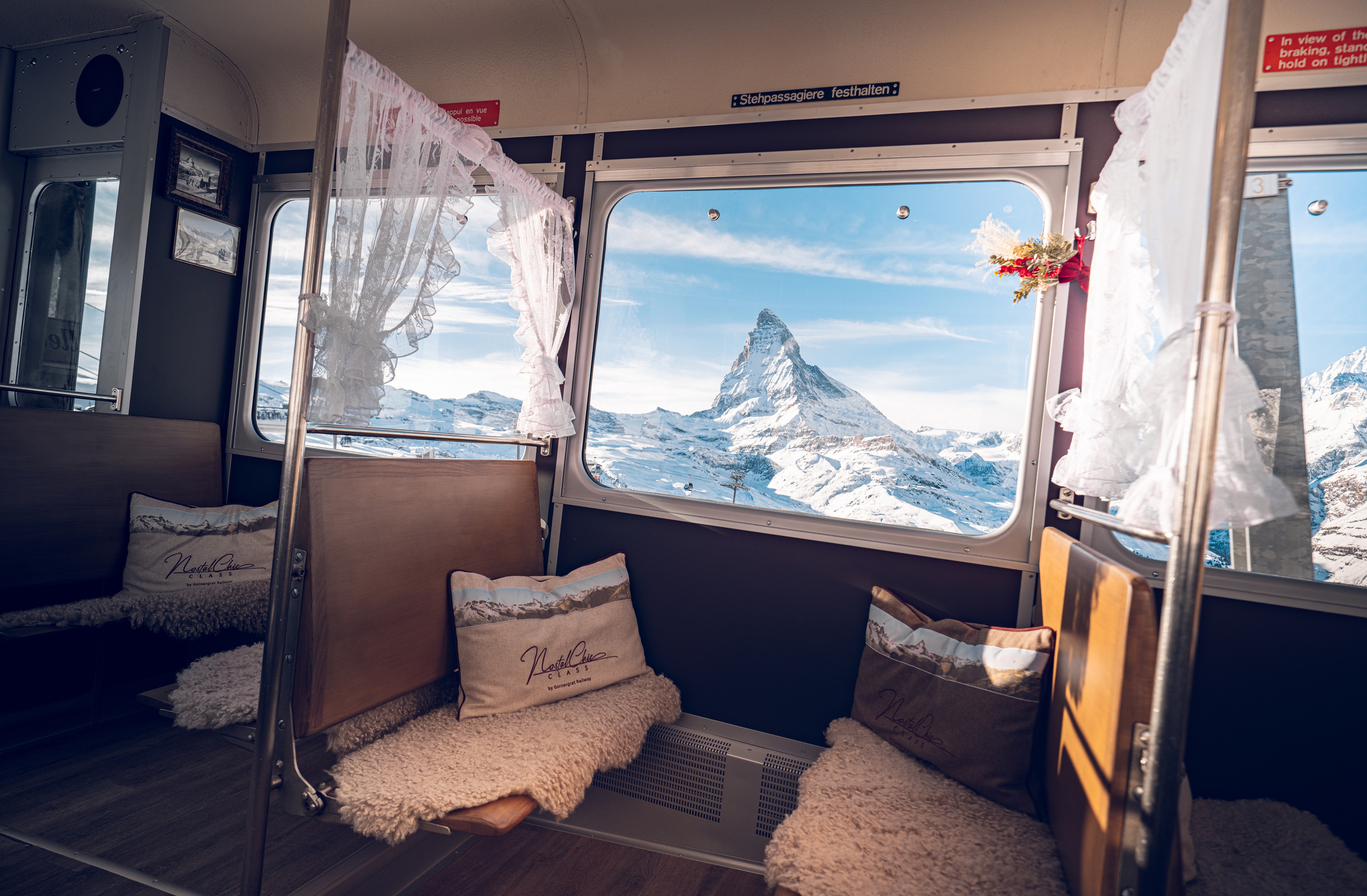 The NostalChic carriage, decorated with cushions and curtains, offers a warm atmosphere. Through the window, the snow-covered Matterhorn is revealed under a blue sky.