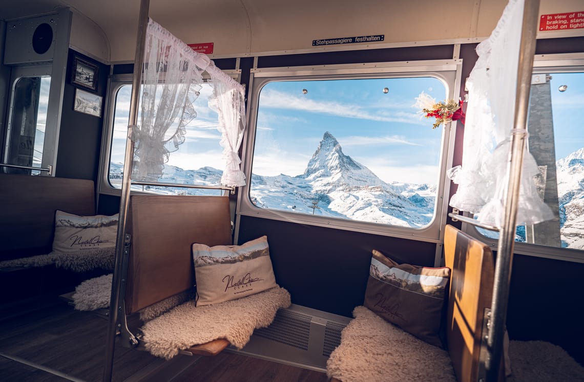 NostalChic Class, Innenansicht, Dekoration und Blick aus dem Fenster aufs Matterhorn Der mit Kissen und Vorhängen geschmückte Waggon des NostalChic bietet eine gemütliche Atmosphäre. Aus dem Fenster zeigt sich das schneebedeckte Matterhorn unter einem blauen Himmel
